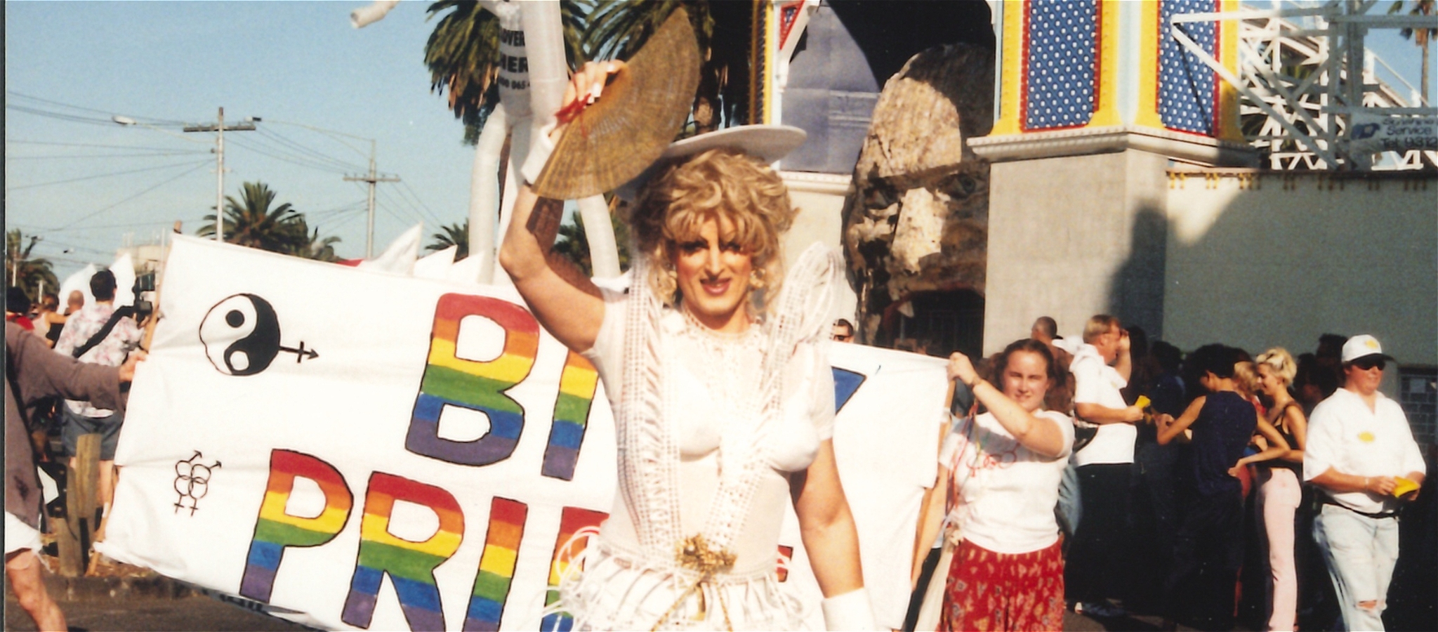 A person in drag wearing a white dress and holding a fan stands in front of a pride banner.