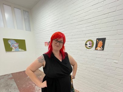 Liz Reed standing in a gallery space or art workshop with hands on hips wearing a black dress and shoes
