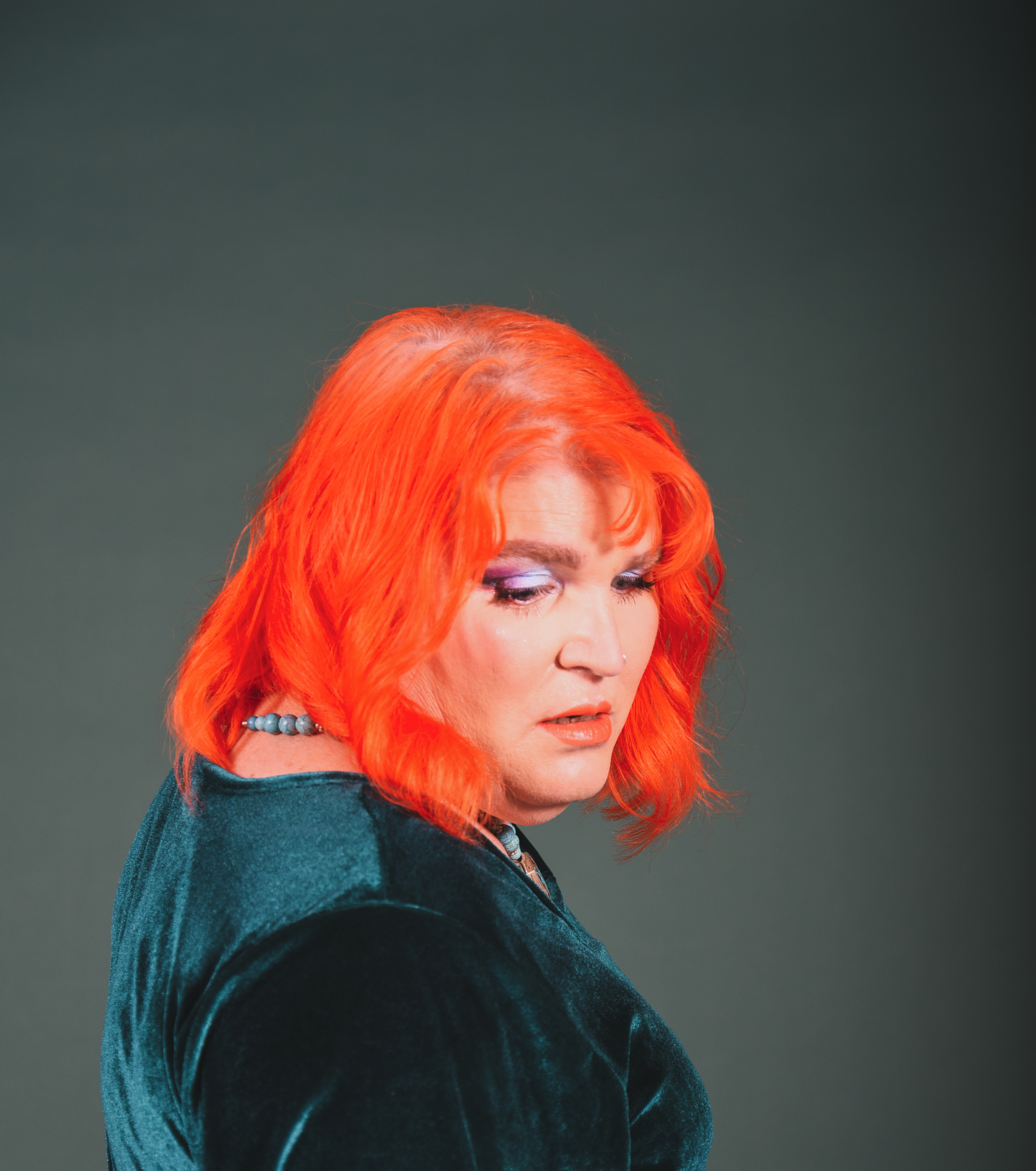 Saint Ergo in a red wig and velvet dress looking downwards