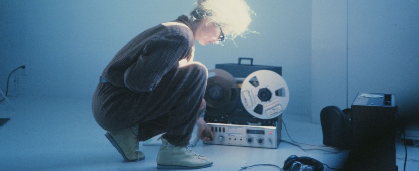 A person with long white hair kneeling down to adjust a large reel tape recorder