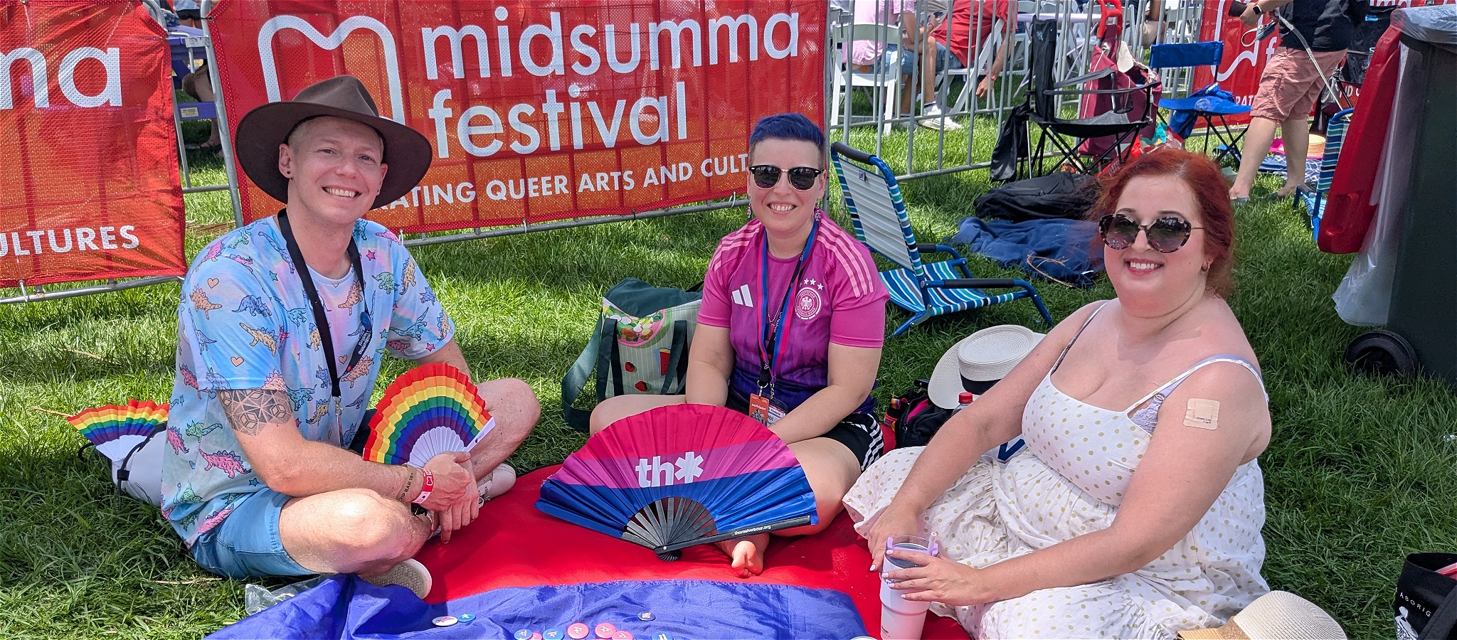 Three smiling people seated around a picnic blanket with bisexual flag and rainbow flag-themed fans in the foreground.