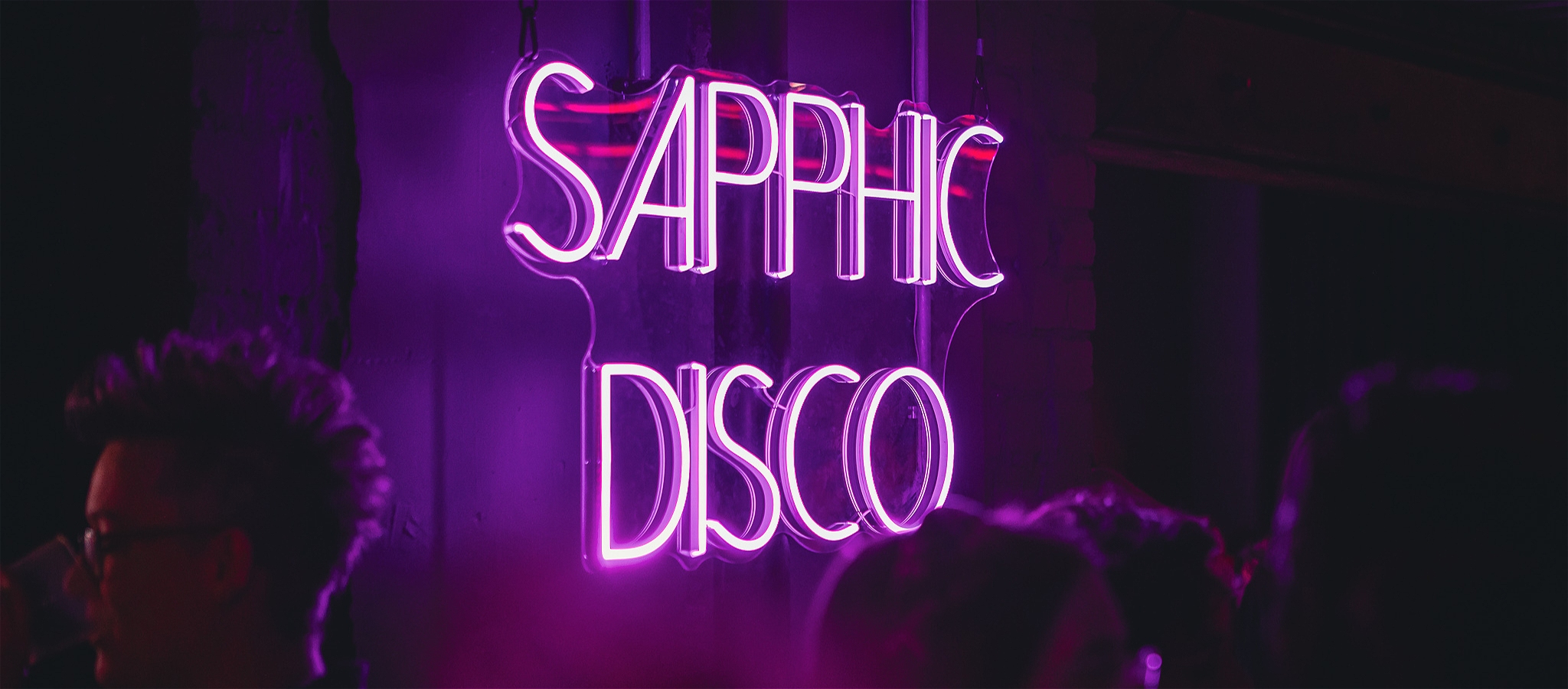 An image of a neon sign on the wall in a club, with the tops of people's heads showing below it
