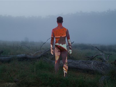 A lone Indigenous man stands naked against the horizon in a misty landscape among fallen tree trunks, their body patterned with coloured light