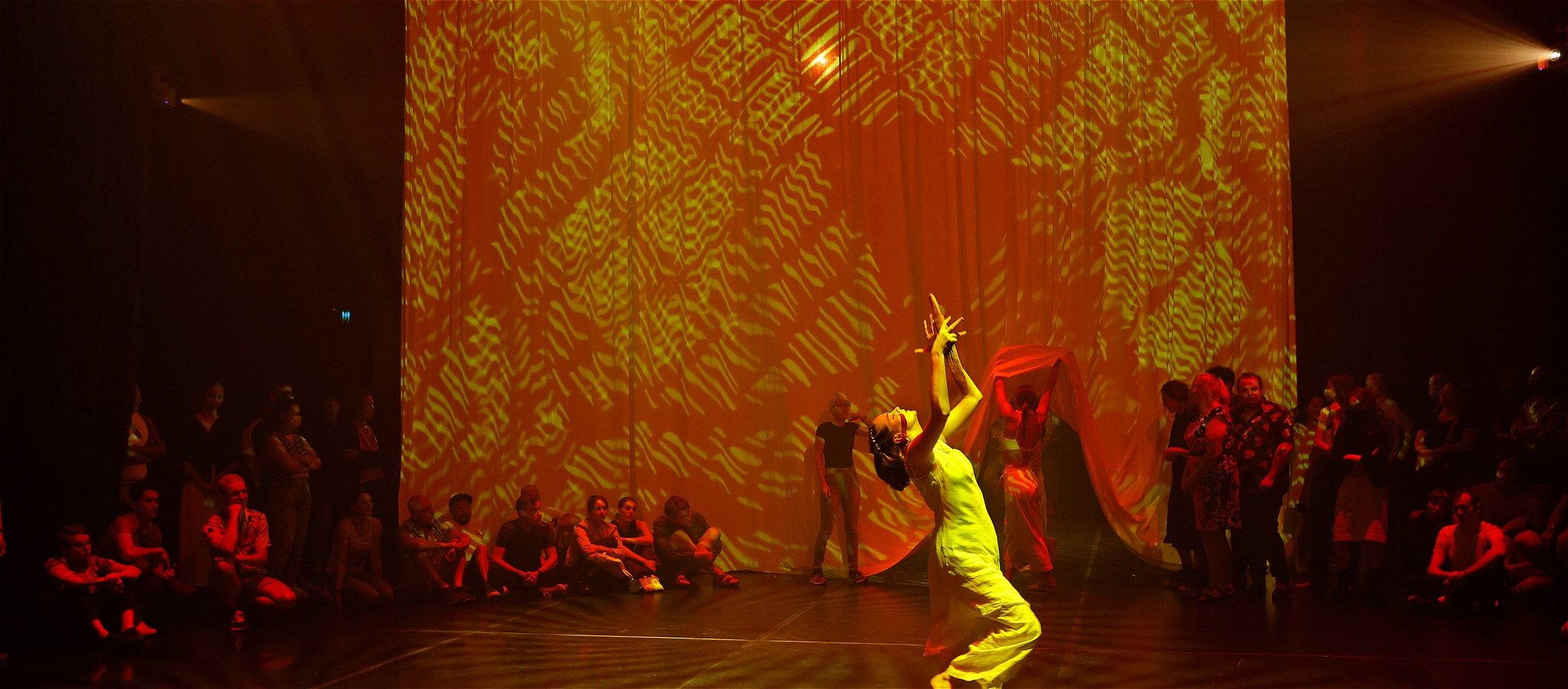 Blindfolded dancer in a yellow dress looking skywards with people sitting on the floor looking on