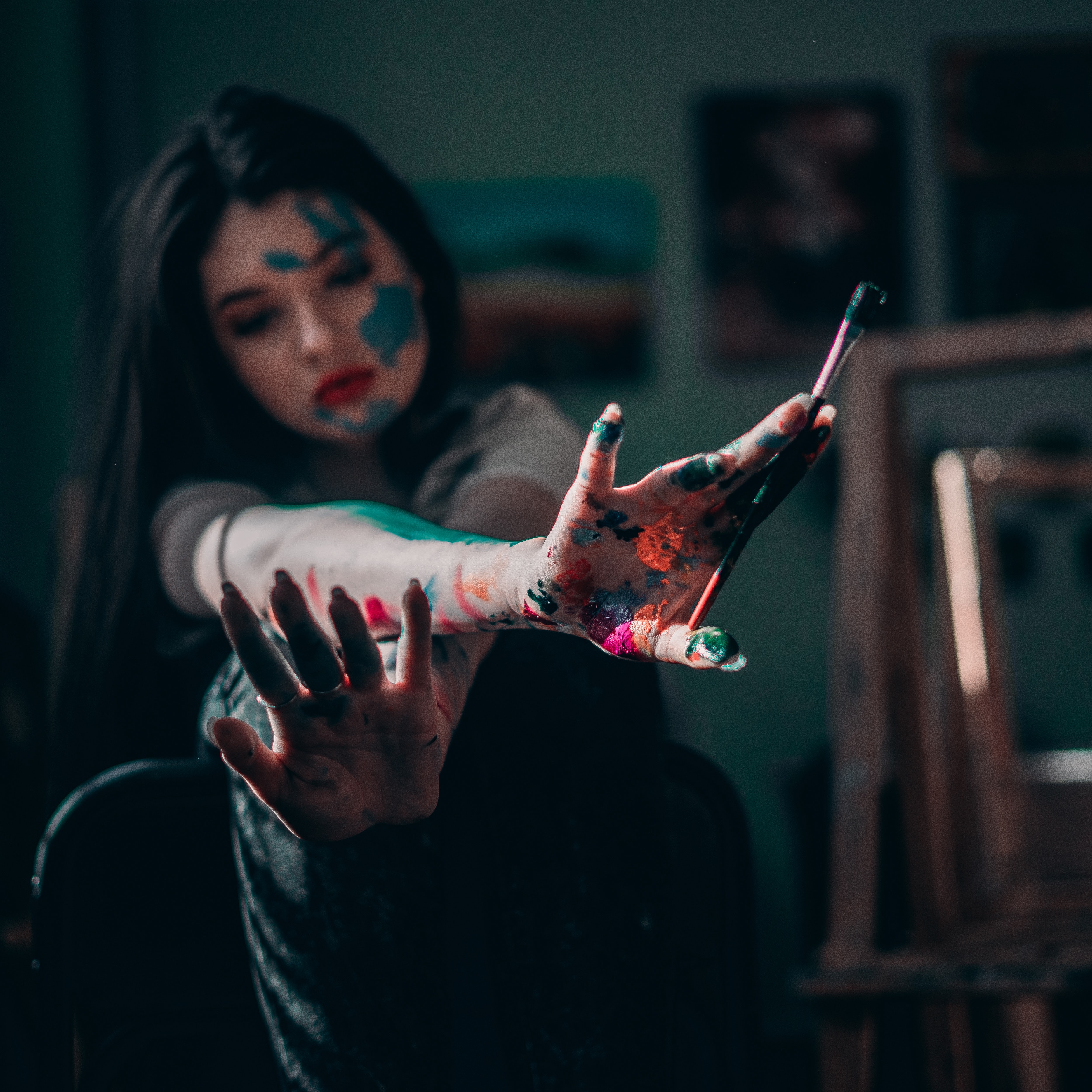 Girl holding a paint brush, with a lot of paint of different colours splashed all over her