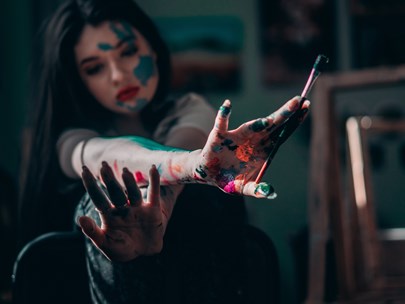 Girl holding a paint brush, with a lot of paint of different colours splashed all over her