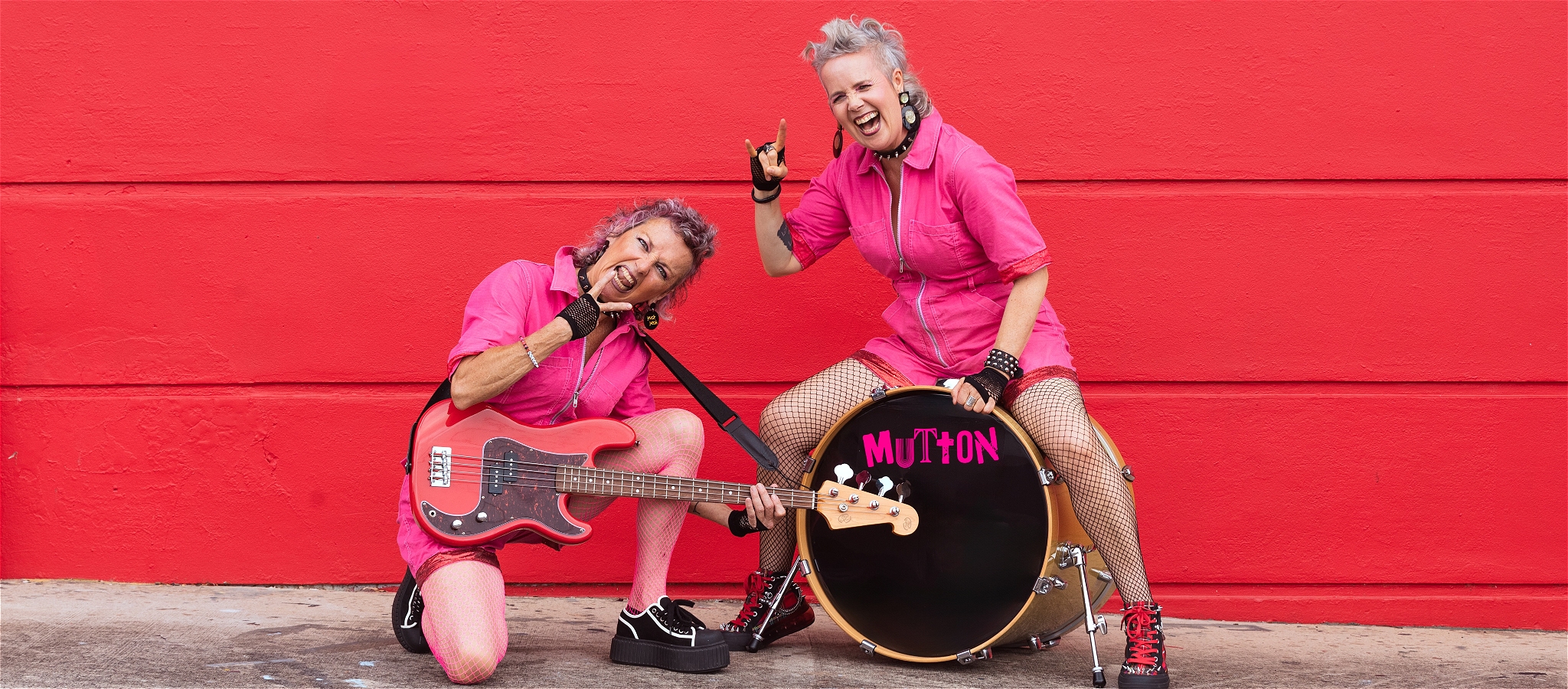 Two women in hot pink short overalls posing with a bass and a drum kit, smiling