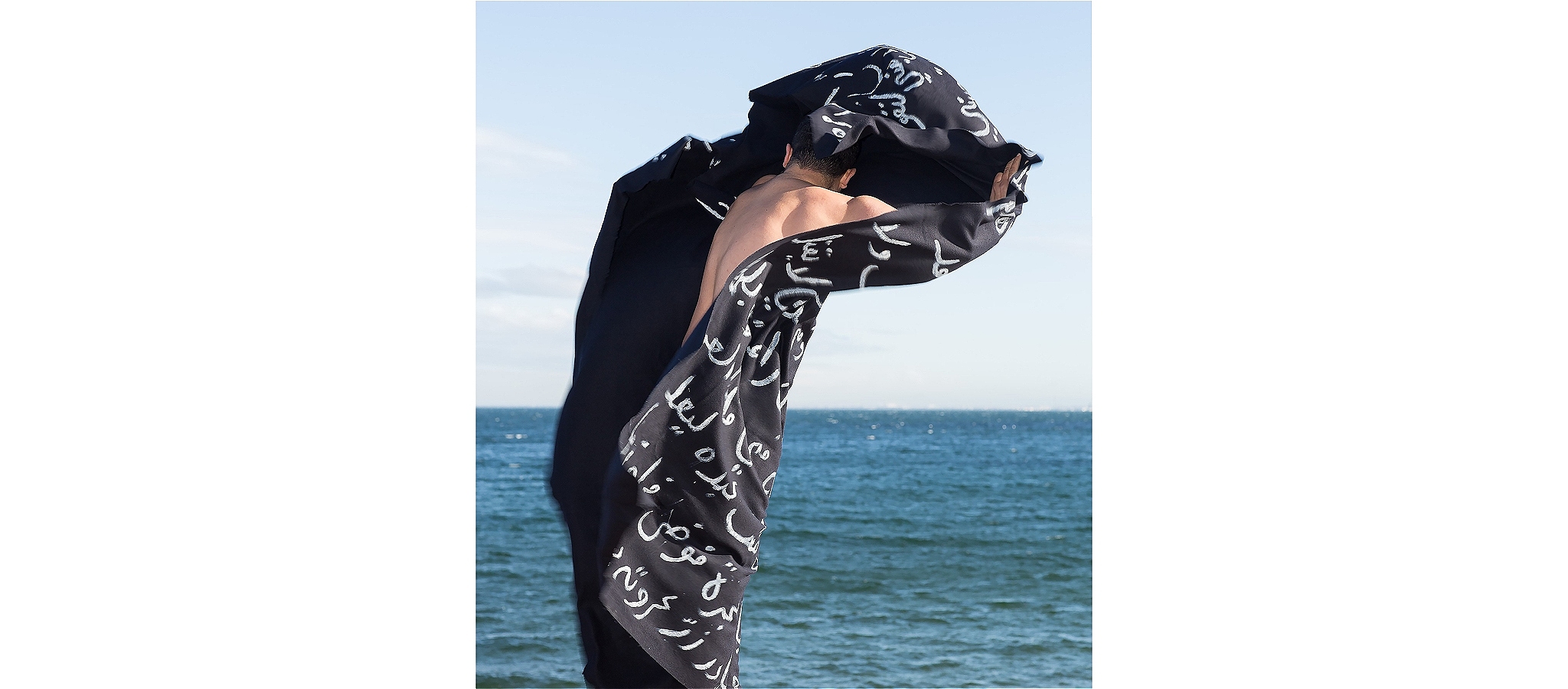 A figure standing in front of the sea, wrapped in a black shroud, covered in white Arabic text