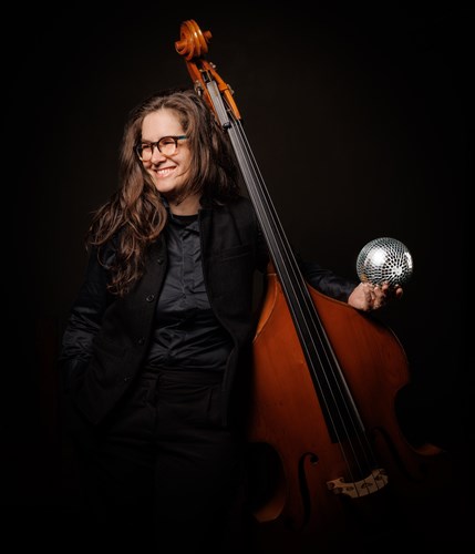 Miranda Hill poses with a disco ball in her left hand whilst a double bass leans against her shoulder. She is wearing glasses and dressed in a black suit against a black background.