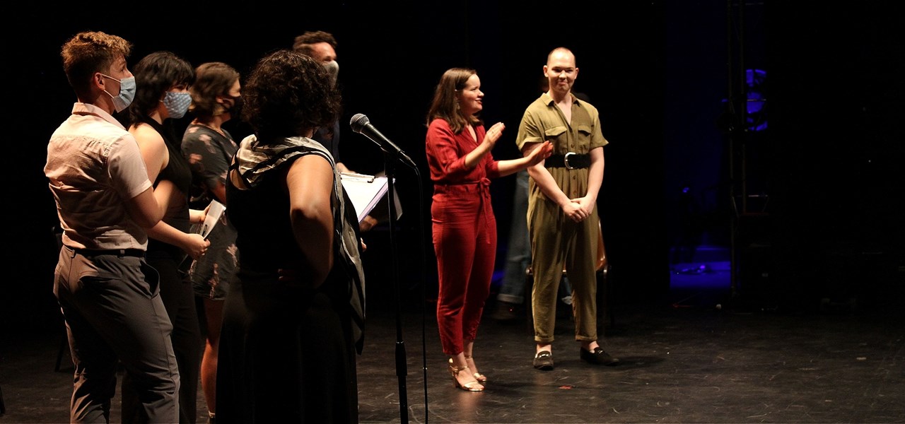 4 actors and 2 playwrights address an audience onstage in a black box theatre; the MC is in the foreground