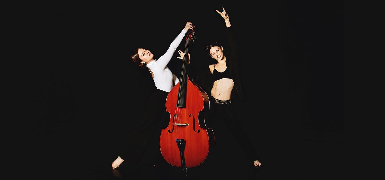Cello in centre with dancer in white to the left with foot outstretched and dancer in black pants, crop and jacket to the right doing the 'rock' symbo