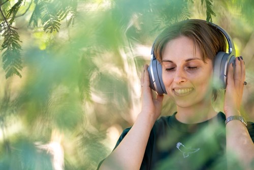 Caitlin is shown sitting in nature wearing headphones, smiling with her eyes closed. She has a septum piercing and is wearing a black shirt. 