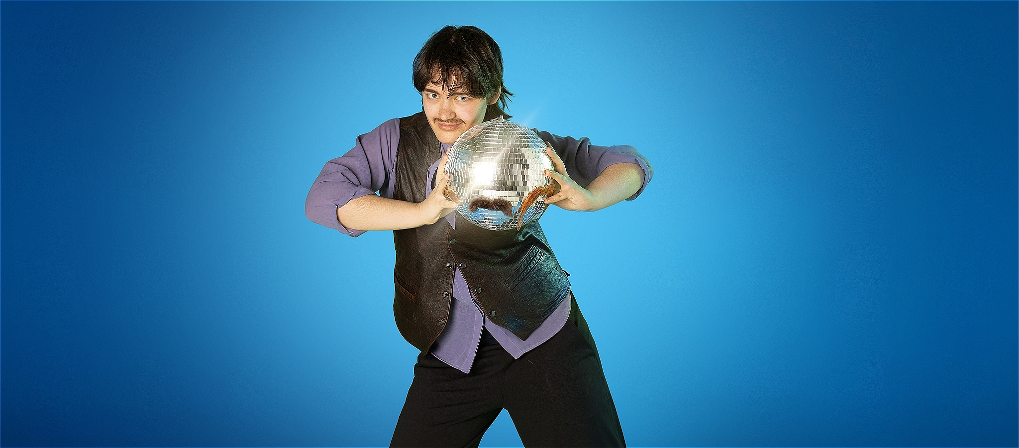 A person dressed in mauve shirt and black vest holds a mirror ball while looking towards the camera