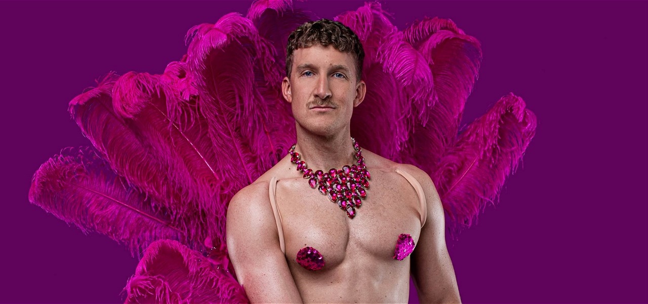 Ziegfeld Boy looks at the camera wearing a hot pink feather backpack, nipple pasties and a jewelled necklace.