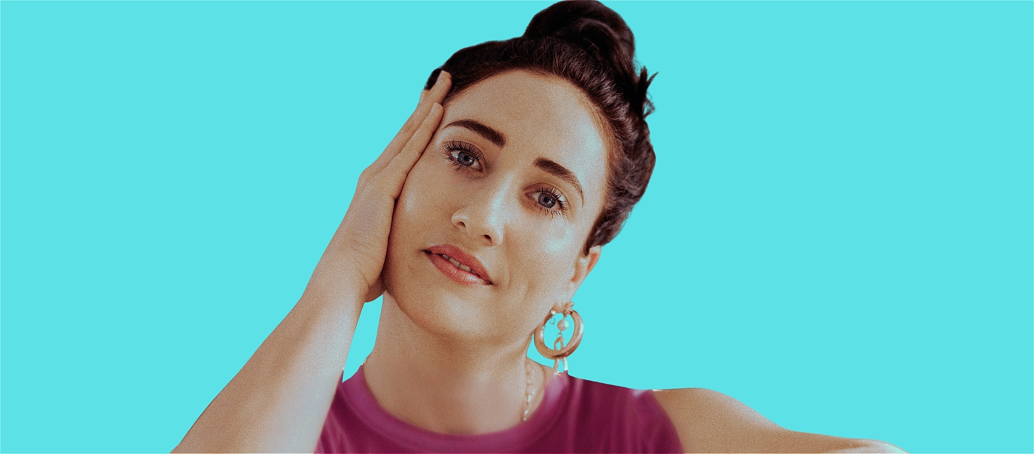 Brunette woman, hair in bun, wearing pink tank top, leaning on her hand, turquoise background
