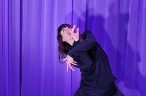 Headshot of Alice on stage with mauve curtains behind, dressed in a black dress and in a 