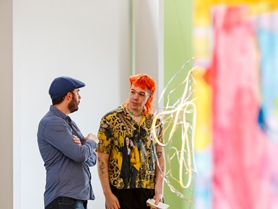 Two people standing together chatting in a gallery space. There is abstract art in front of them, perhaps viewed through a window
