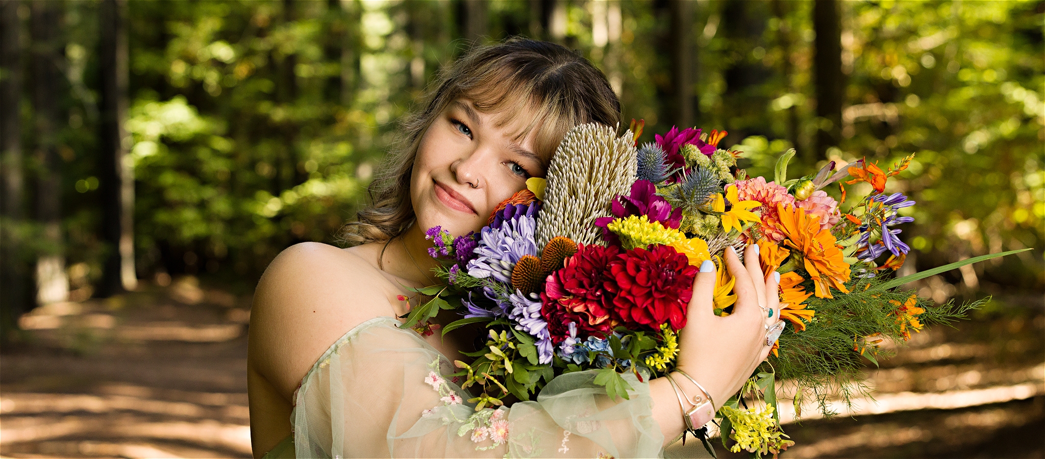 A vibrant portrait of queer, female singer/songwriter Mieke Bosland standing in a forest, wearing a floaty teal dress and holding a rainbow bouquet.