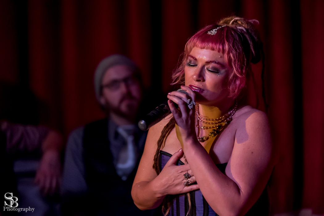 Woman singing in a soulful way, eyes closed. A bearded man is behind her, a little out of focus