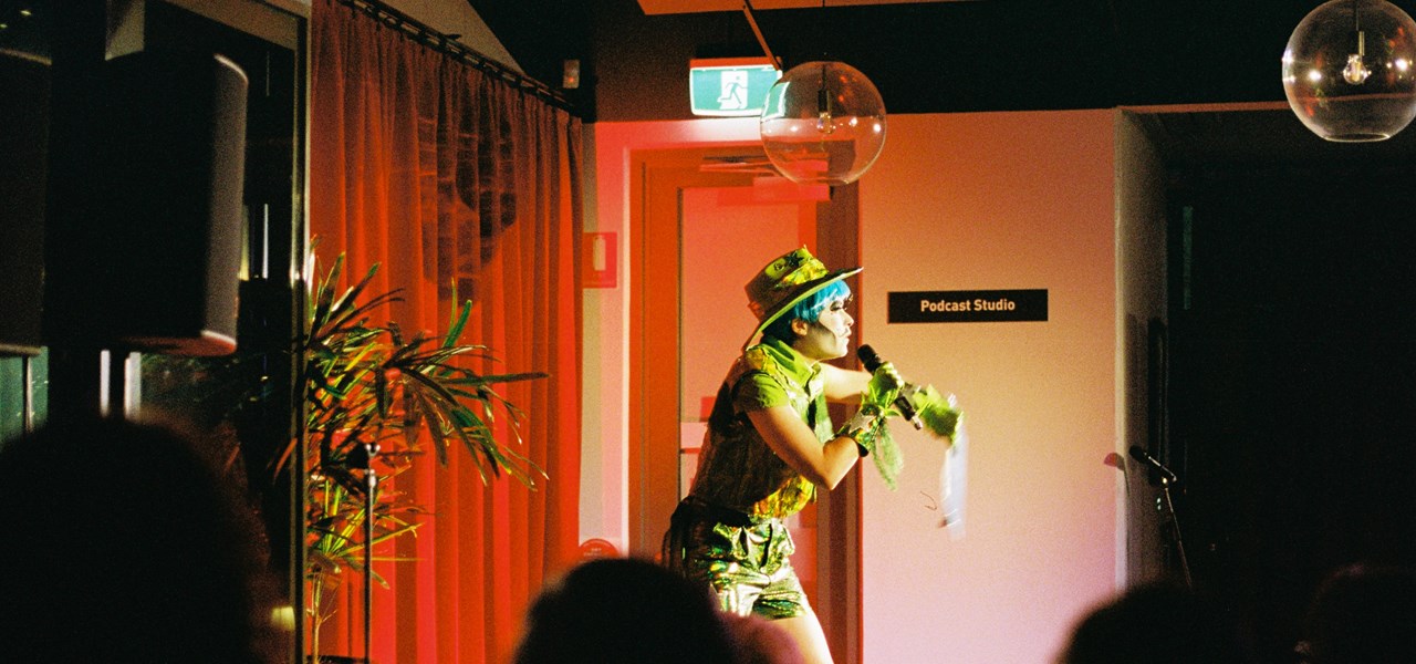 A person in a blue wig, wearing a green hat, shirt and shorts holds a microphone in front of a red curtain. 
