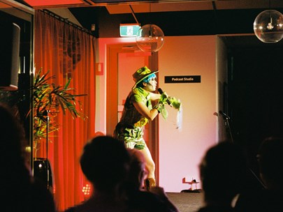 A person in a blue wig, wearing a green hat, shirt and shorts holds a microphone in front of a red curtain. 