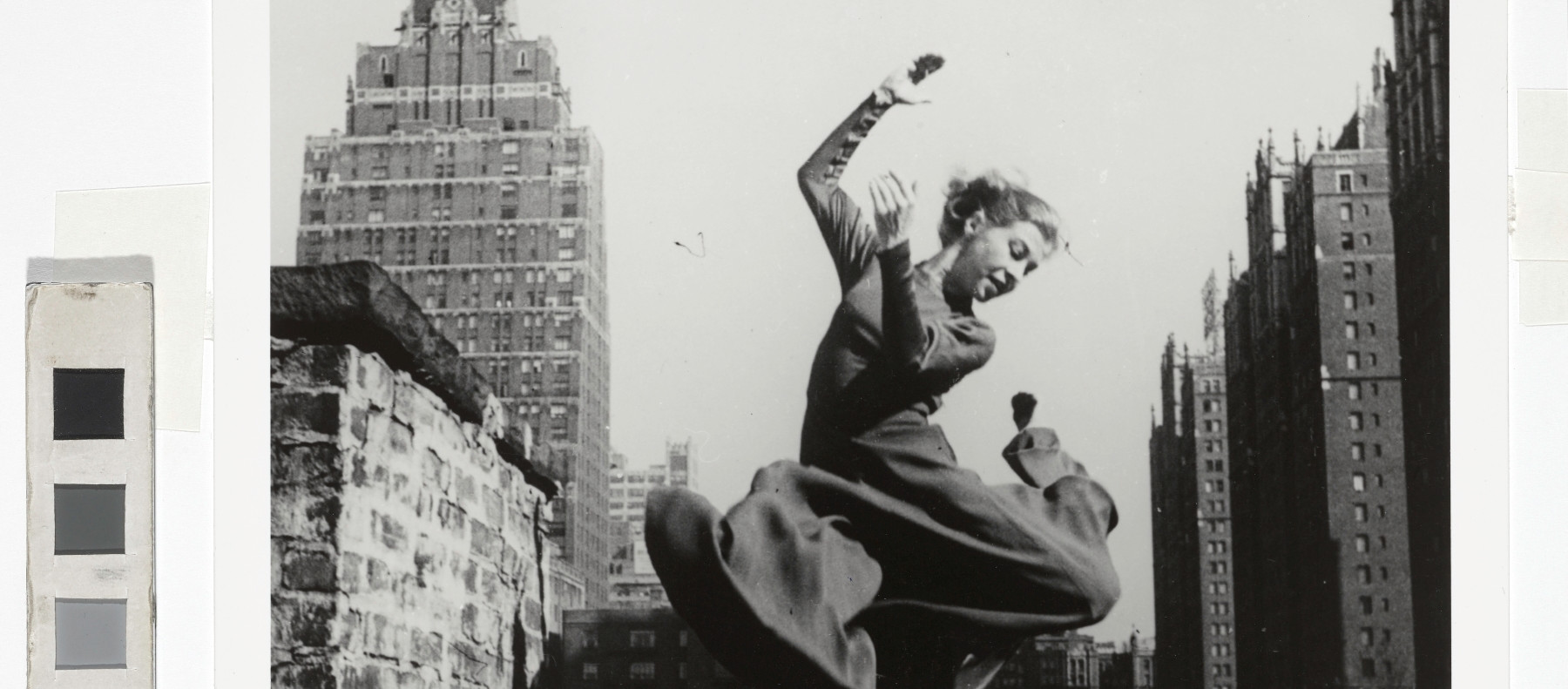 A black and white photograph of a woman wearing a dark coloured dress, jumping with joy