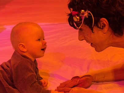 A baby and an adult in a sparkly costume crawl towards each other smiling in a space with red and pink lighting.
