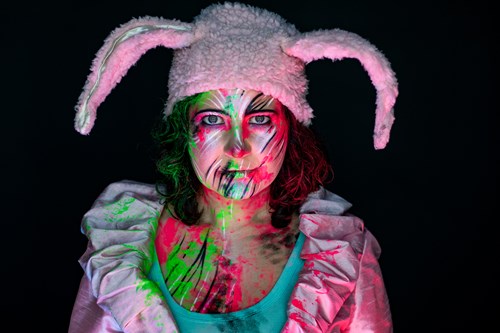 Jessie gazes intensely into the camera, while wearing brightly coloured make-up and a pink, plush hat with two ears. 