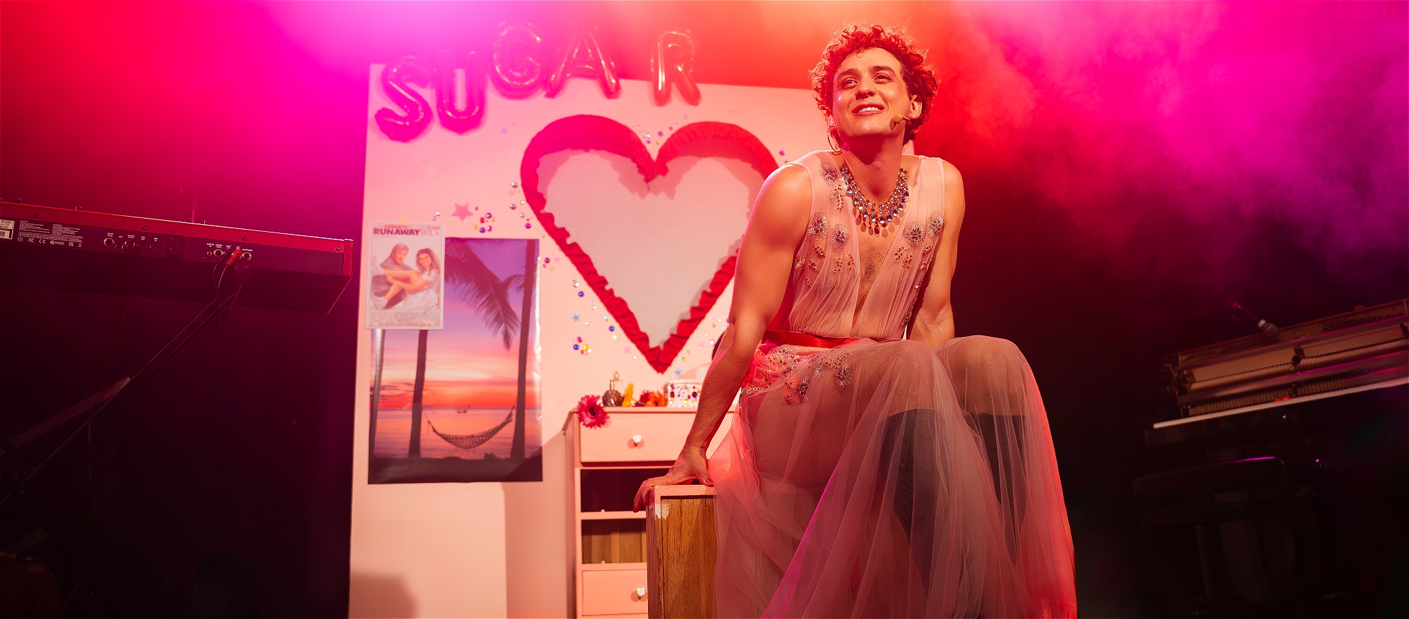 a person in a sheer pink dress sits infront of a pink bedroom scene. the words sugar are written in red balloons with a heart cut out in the wall.
