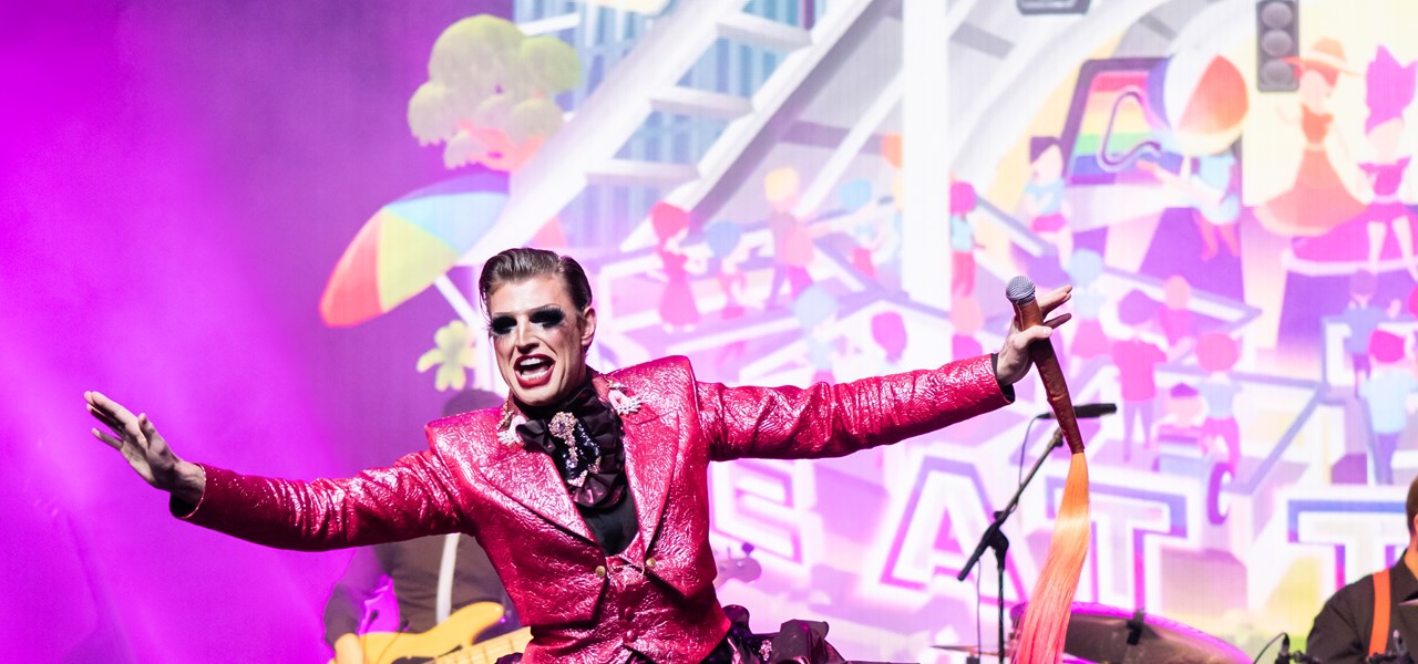 Reuben Kaye on stage with a set of drums behind them and the Midsumma 2021 theme in the background