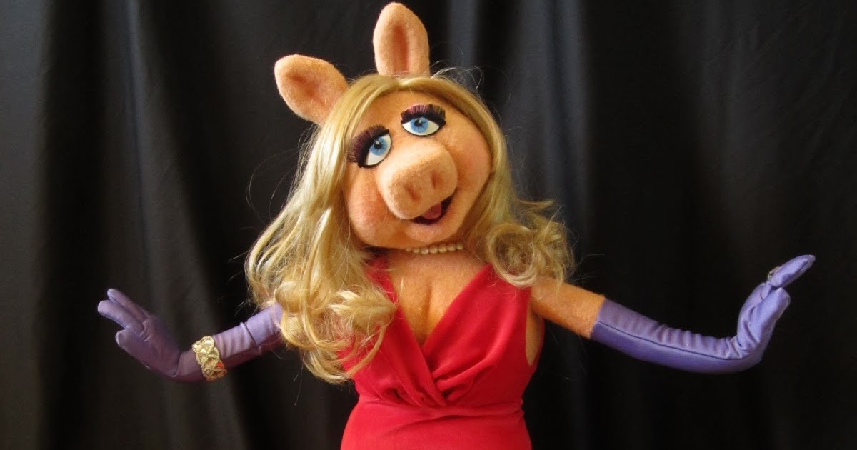 Performer wearing a Miss Piggy mask, red dress and mauve gloves in front of a black curtain