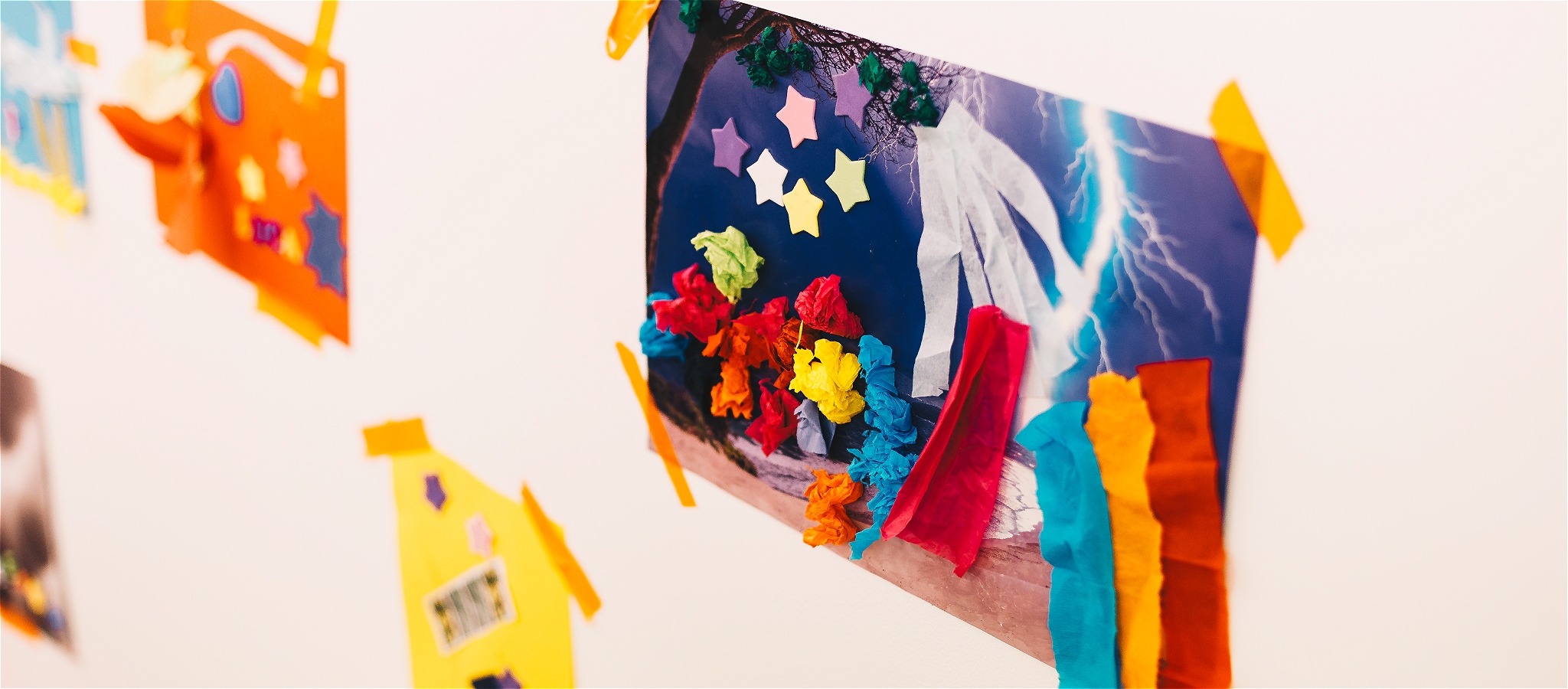 An artwork of a colourful collage containing blue, red, yellow, orange and green scrap paper displayed on a white wall.