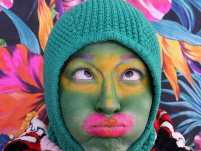 Person wearing a green knitted beanie and colourful top surrounded by painted flowers. They are looking cross-eyed.