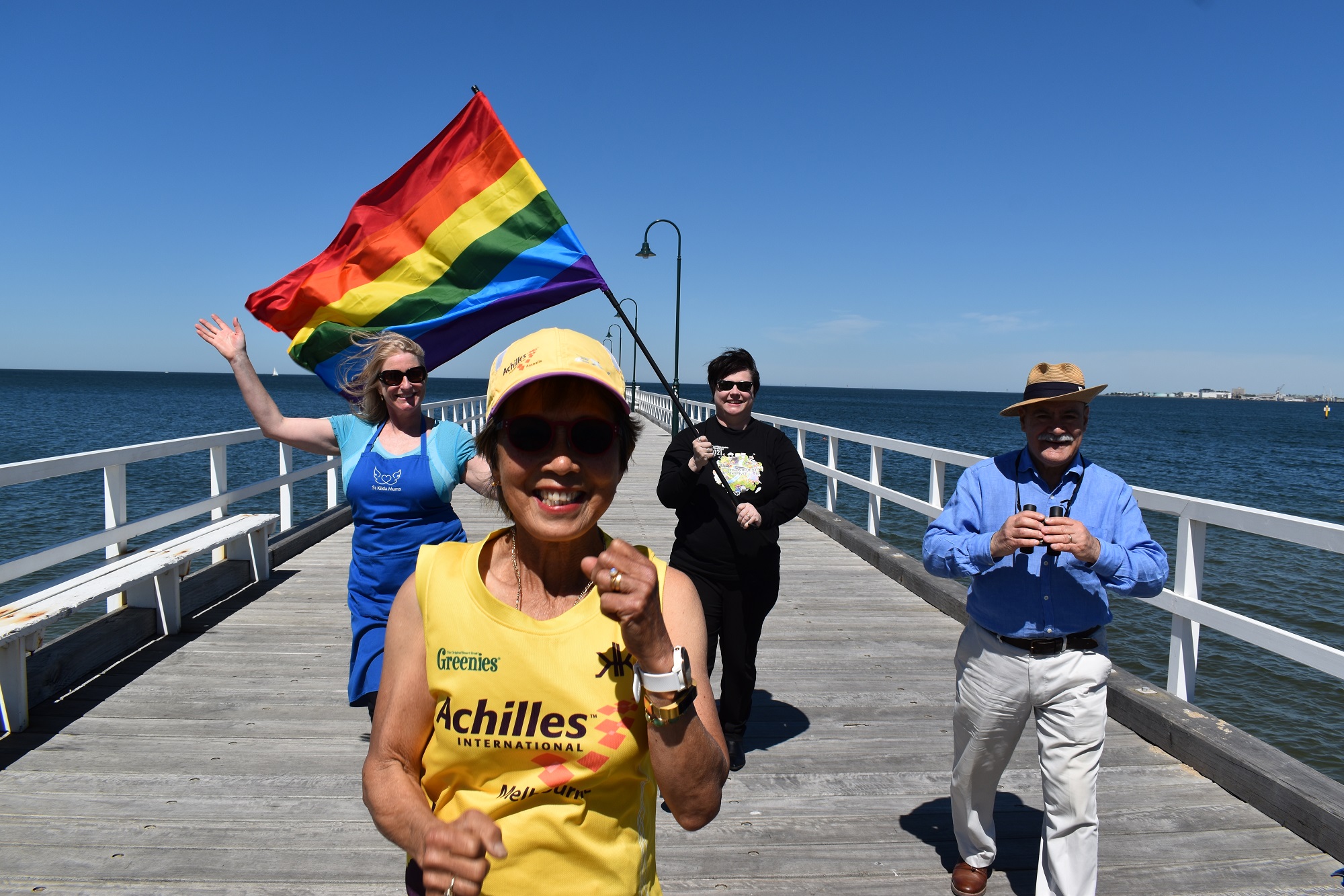 Four people walking on a jetty, the back two holding a large Pride flag