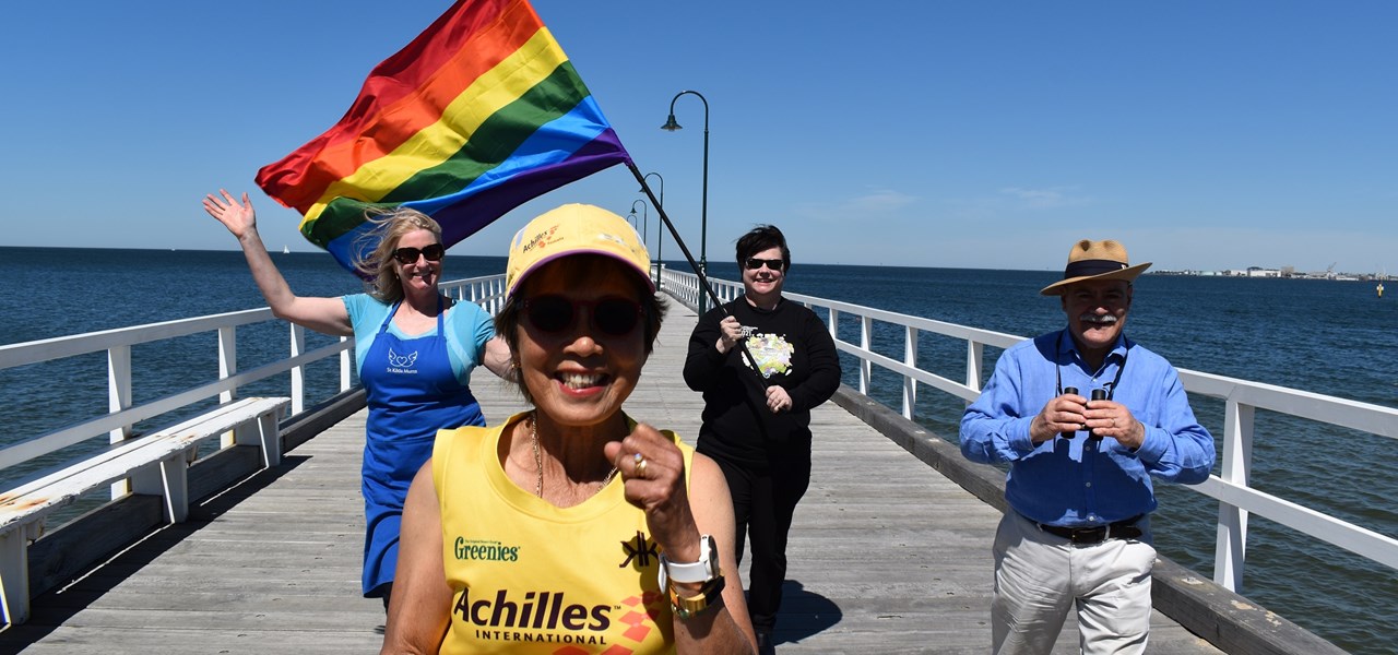 Four people walking on a jetty, the back two holding a large Pride flag