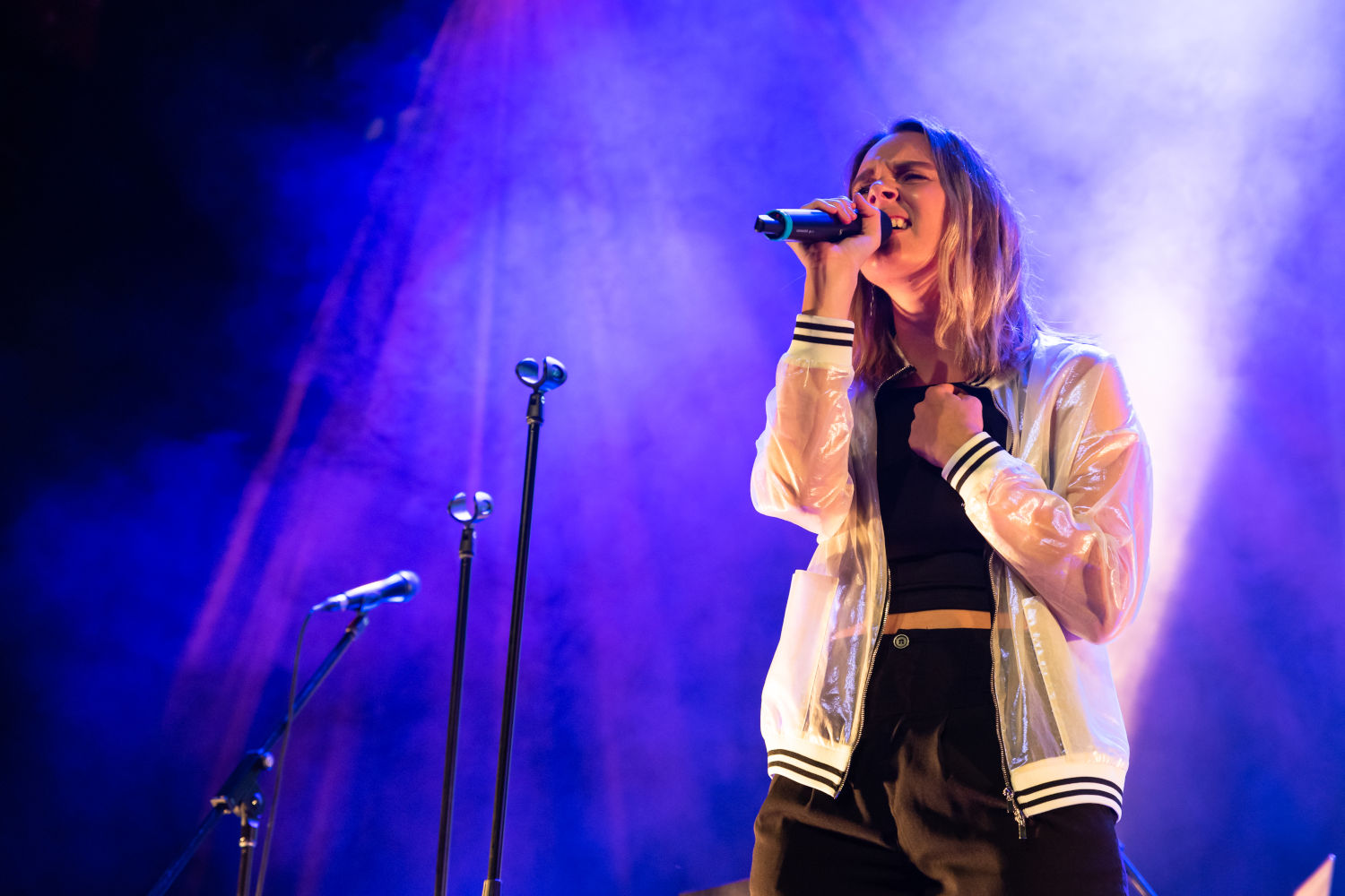 Person on stage singing into a microphone with blue lighting. Several extra unattended microphones