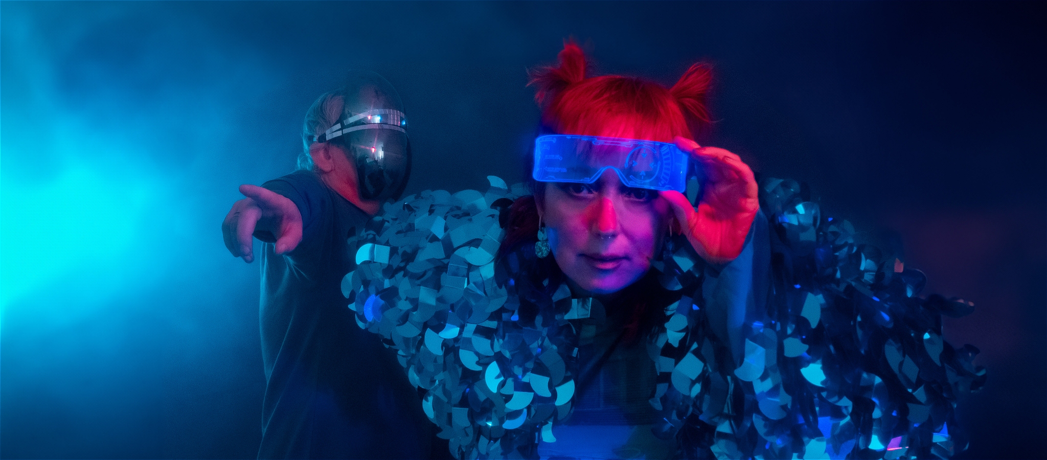 person in robot mask in background pointing person in foreground peering under cyber glasses leaning forwards
