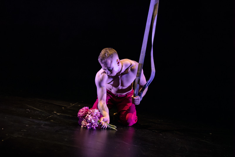 Luca picking up some flowers while holding some ropes - bare chested, looking like they're doing acrobatics