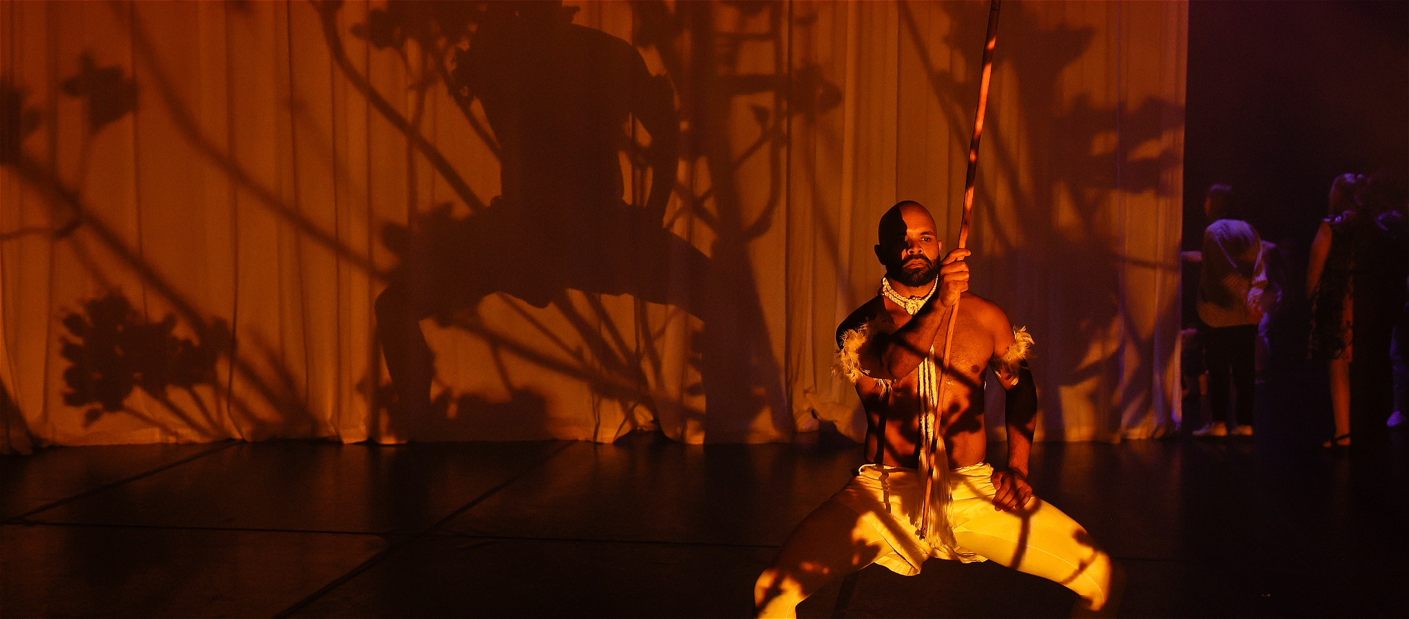 bare-chested dark man on stage holding a stick with lots of shadows projected onto them