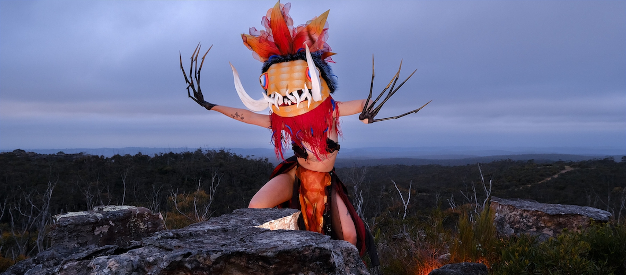 A person dressed in a colourful beast outfit with very long fingernails and tusks coming from their mouth