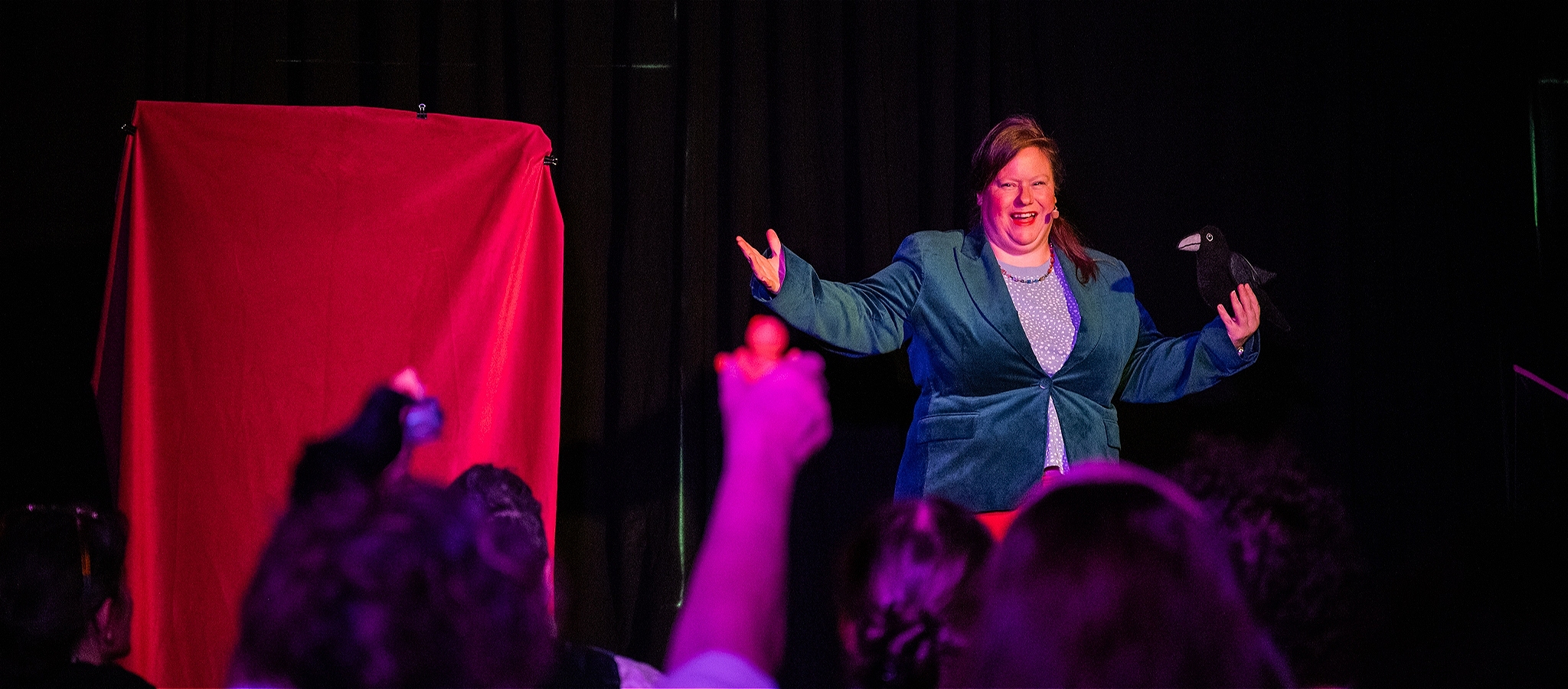 A woman, smiling, on stage, wearing a dark green blazer, blue tshirt and rainbow necklace. Holding a plush raven.