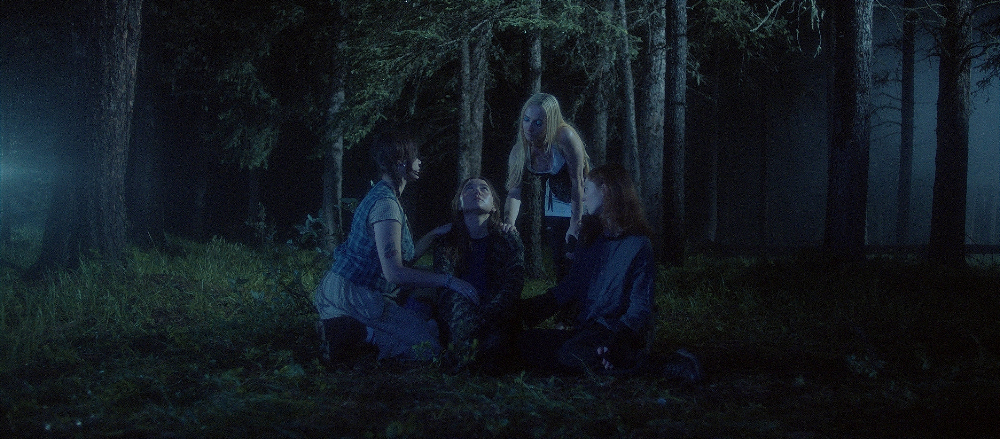 Four Femme presenting people in a huddle sat on the floor of a dark and scary looking forest.