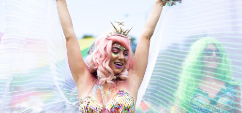 Person in a pink wig, wearing a crown, arms in air looking very happy - gossamer angel wings behind them