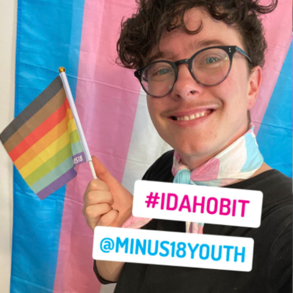 Young person wearing glasses waving a gay flag. Stickers #IDAHOBIT and @MINUS18YOUTH