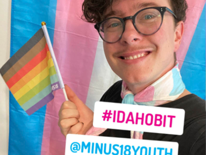 Young person wearing glasses waving a gay flag. Stickers #IDAHOBIT and @MINUS18YOUTH