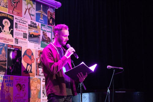 A male-identifying person speaking into a microphone while reading from a booklet. Lots of theatre posters behind them.