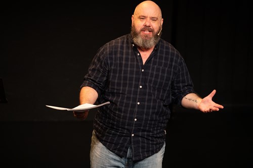 Ben is shown on stage wearing a microphone headset and a checkered shirt, holding a book of papers in one hand. 