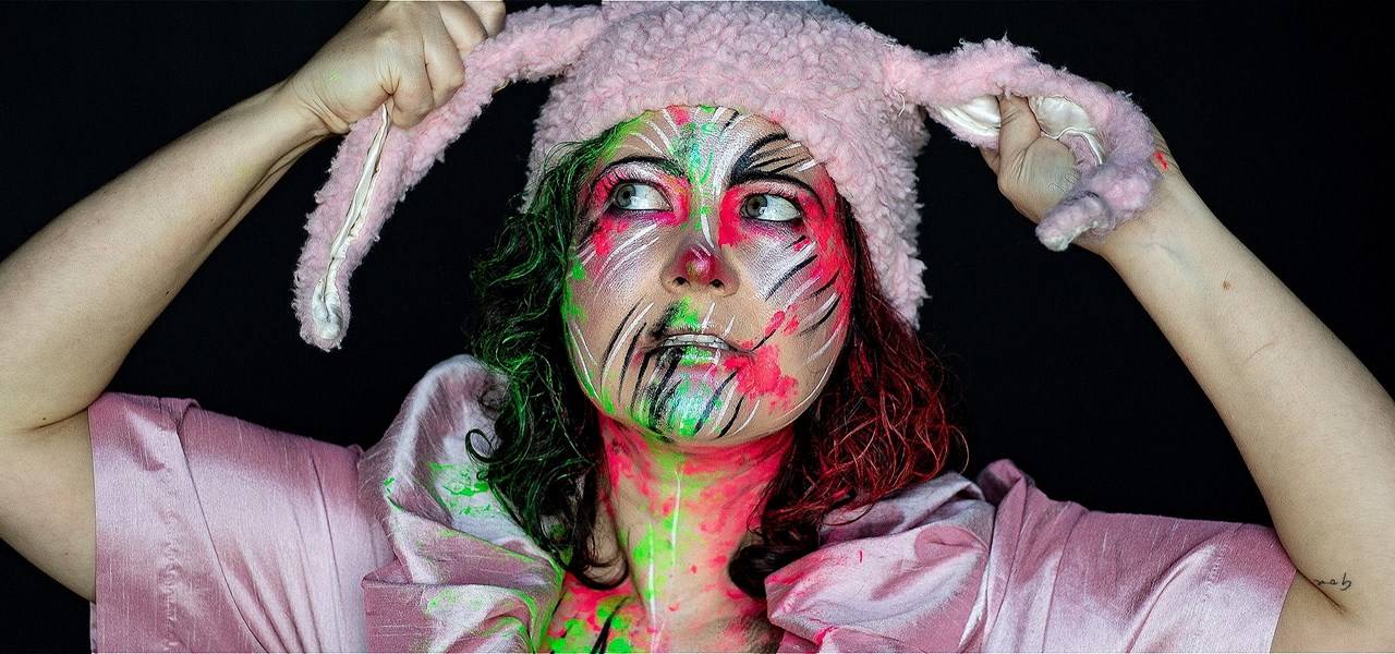 A woman dressed all in pink with a painted face is wearing fuzzy pink bunny ears, which she is gripping in her hands.