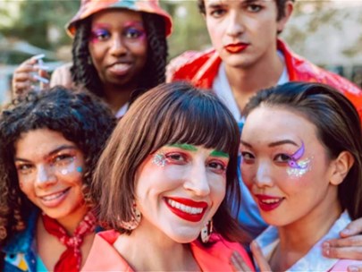 Six people in brightly coloured clothes and sparkly eye make-up are huddled together and smiling at the camera.