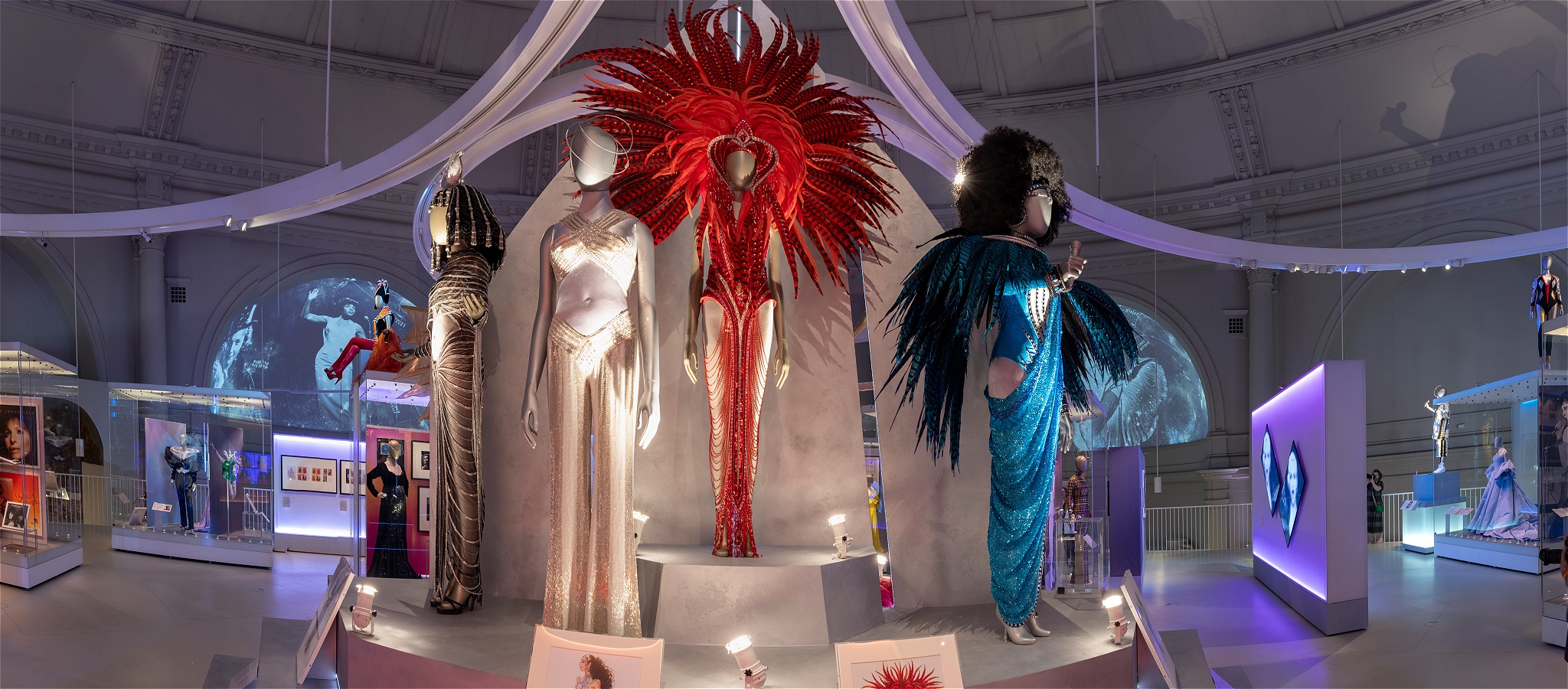 Display of five costumes including a red dress with red feathers and a blue dress with feathers.